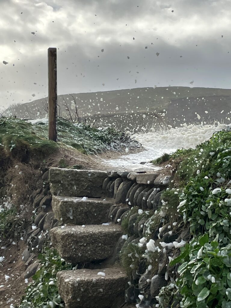 Photo by Caroline Pack of sea foam blowing across a granite style at Gunwalloe