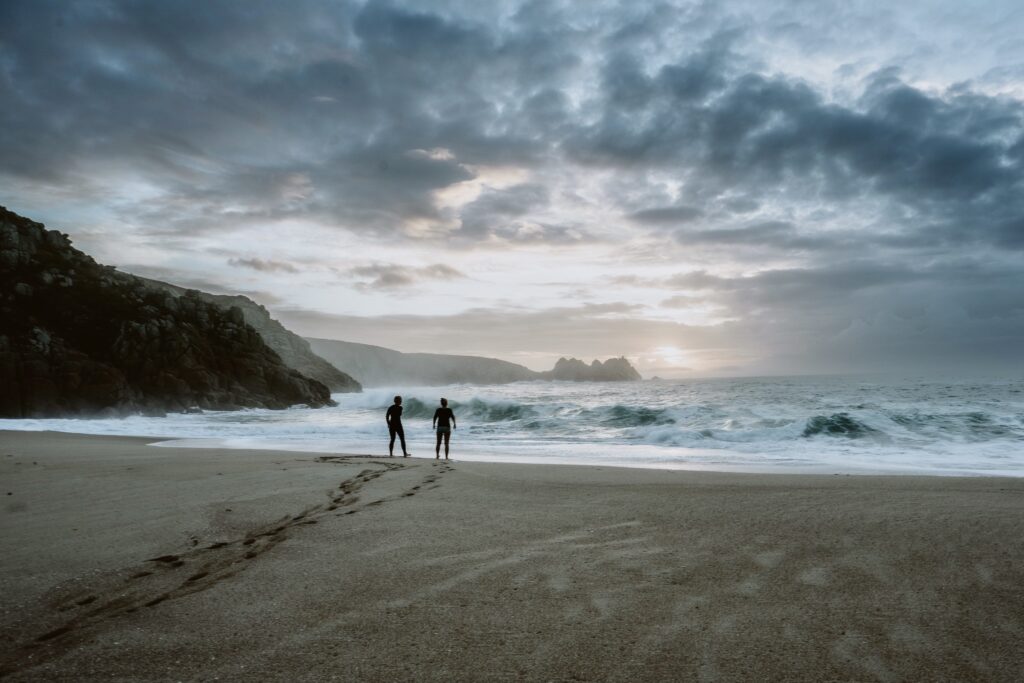 Photo by Jess Feldon of two figures on the beach, looking out to sea at Porthcurno